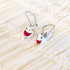 A pair of silver fishhook earrings with small white bird charms featuring red bellies, orange beaks, and black eyes, displayed on a cream-colored ruffled fabric background.