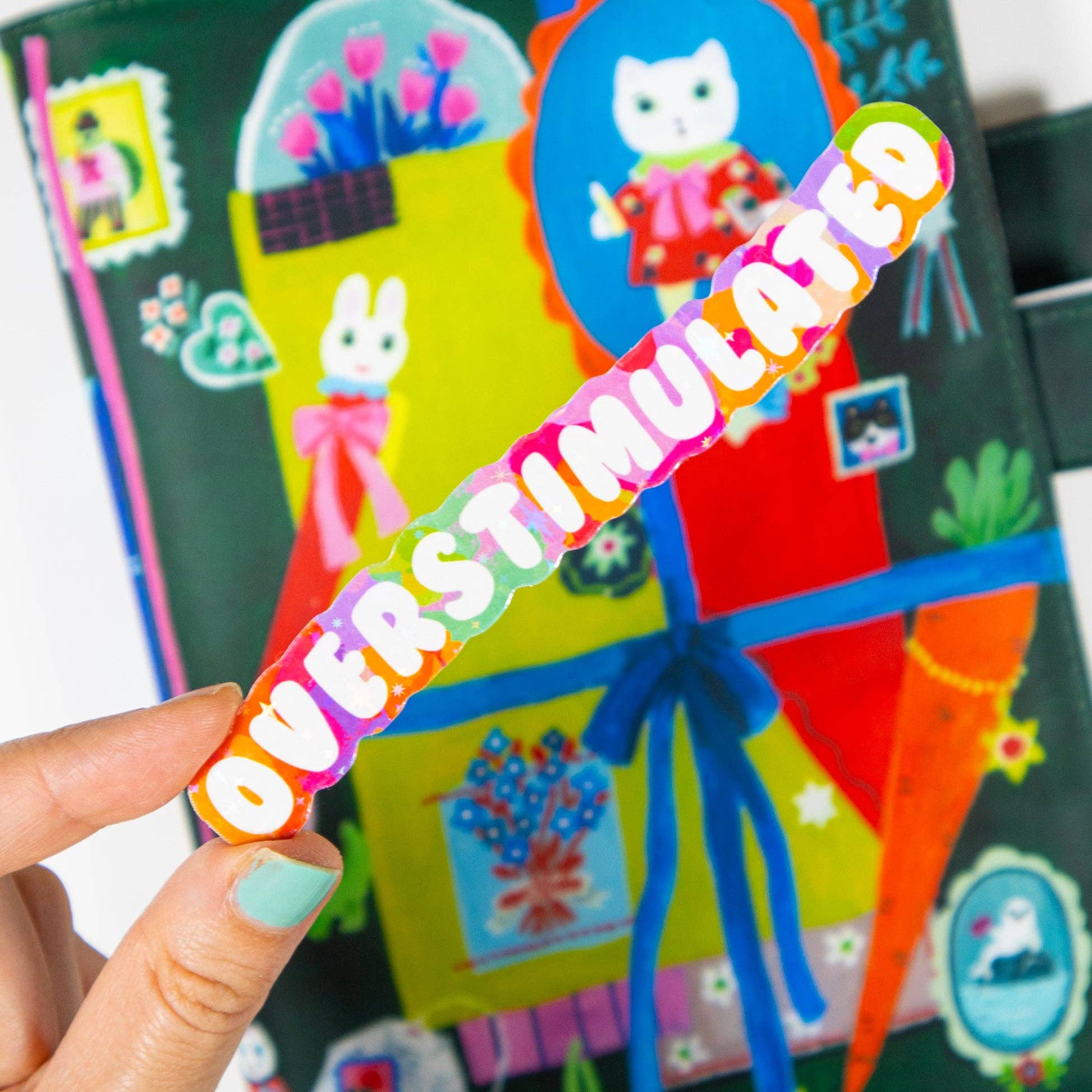 Colorful sticker with 'OVERSTIMULATED' text held by a hand in front of a colorful background