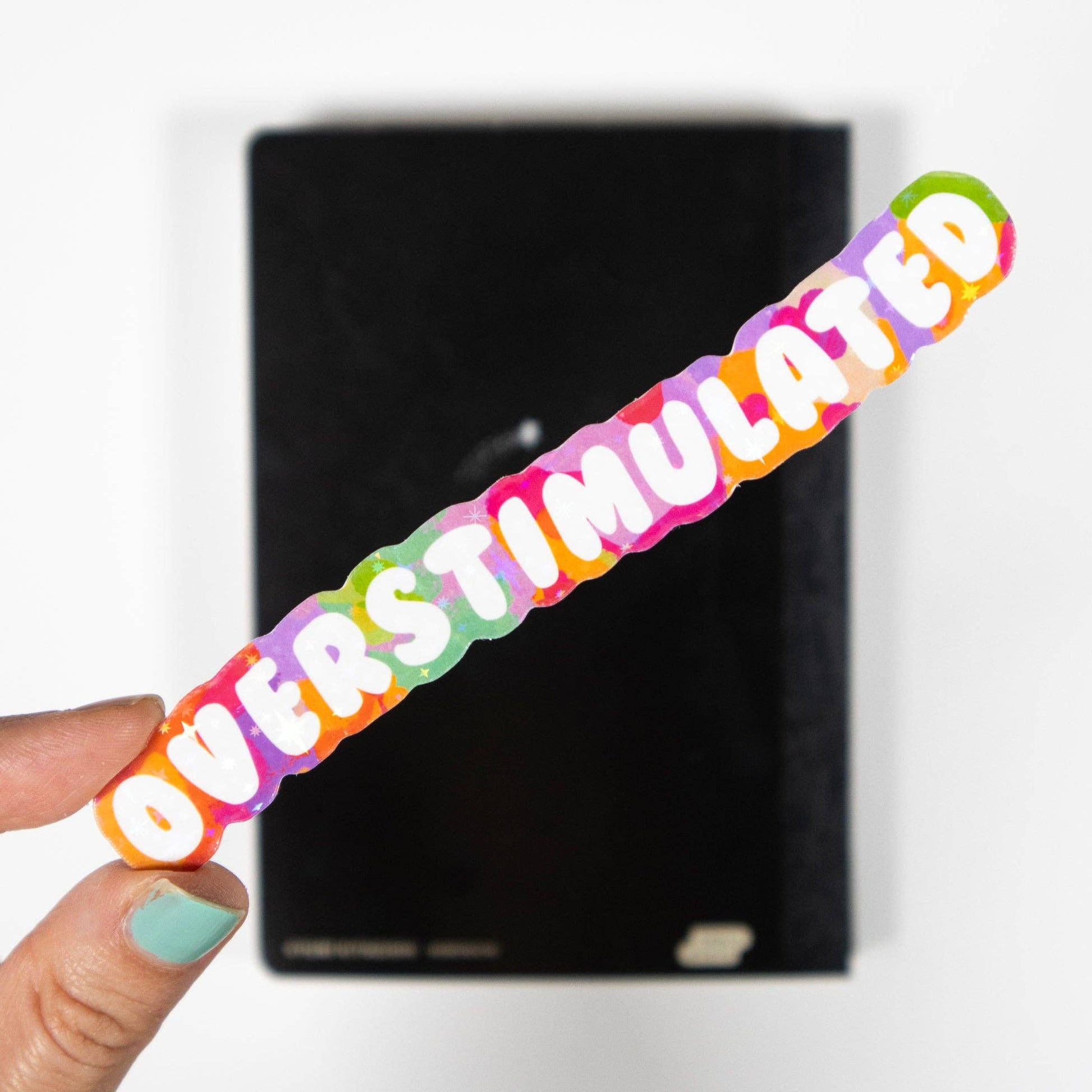 Colorful sticker with 'OVERSTIMULATED' text held by a hand against a black background