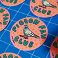 Round pink stickers featuring a pigeon illustration and the text 'PIGEON FAN CLUB' in green, arranged on a blue gridded cutting mat.