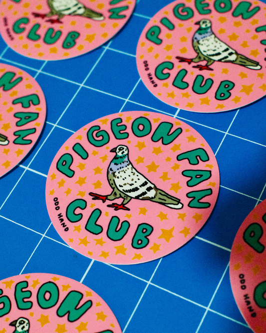 Round pink stickers featuring a pigeon illustration and the text 'PIGEON FAN CLUB' in green, arranged on a blue gridded cutting mat.
