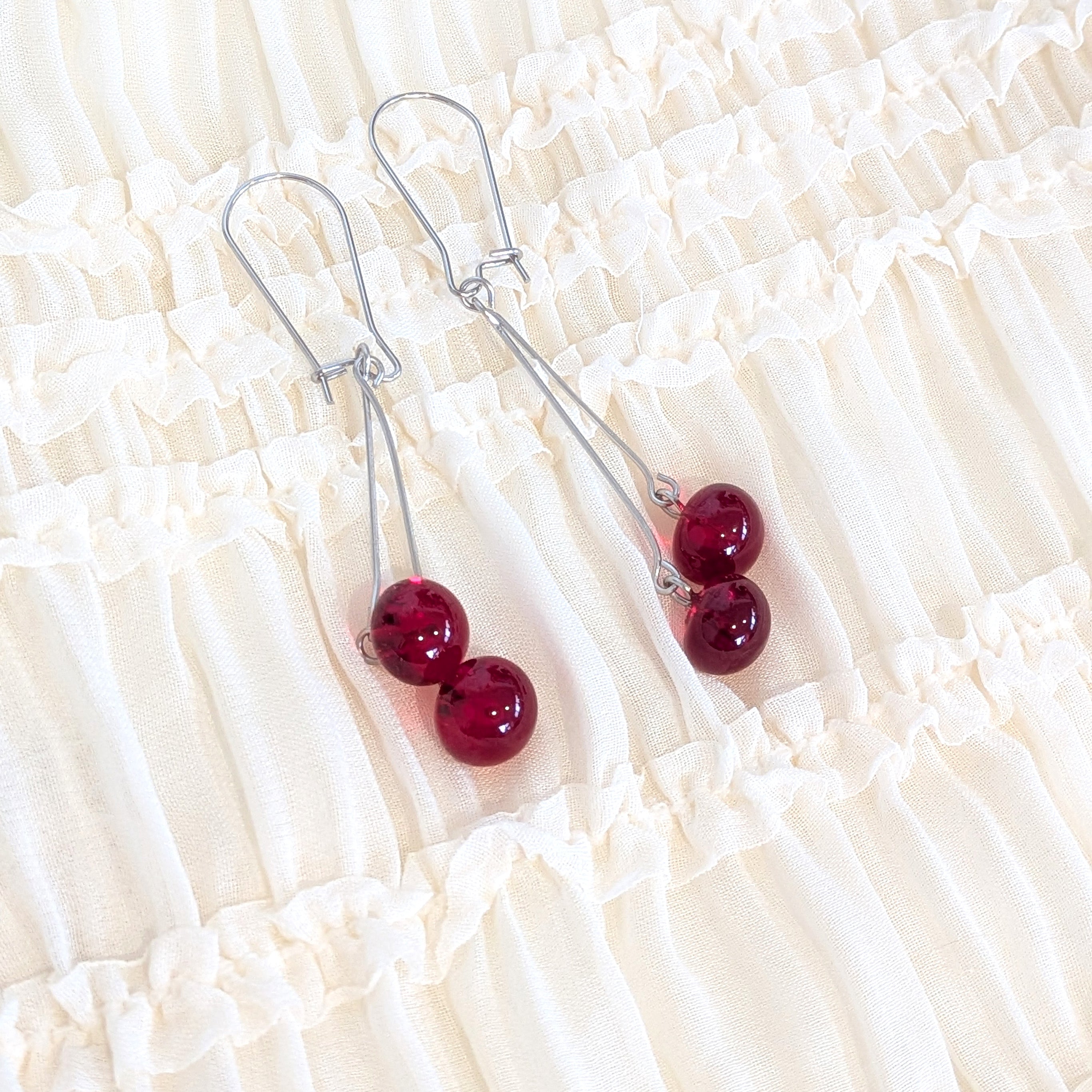 A pair of silver-toned drop earrings with two glossy deep red beads, displayed on a cream-colored ruffled fabric background.