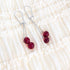 A pair of silver-toned drop earrings with two glossy deep red beads, displayed on a cream-colored ruffled fabric background.