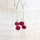 A pair of silver dangle earrings with glossy red bead drops, hanging against a soft white ruffled fabric background.