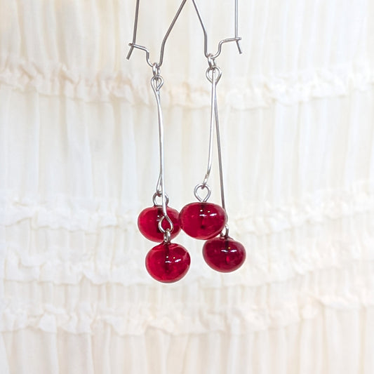 A pair of silver dangle earrings with glossy red bead drops, hanging against a soft white ruffled fabric background.